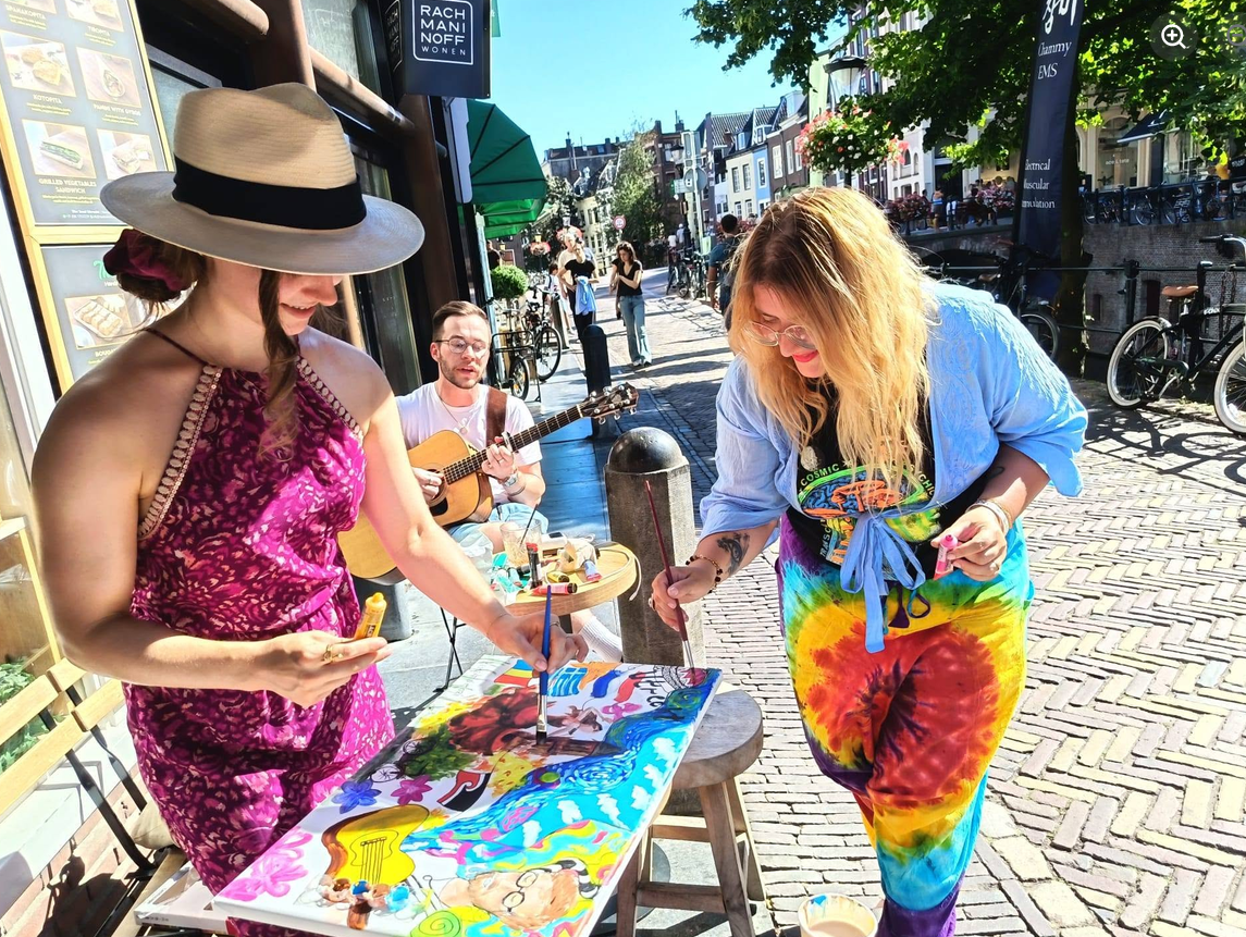 Two people painting a colourful canvas on a street table in the Netherlands while a street musician plays guitar beside them