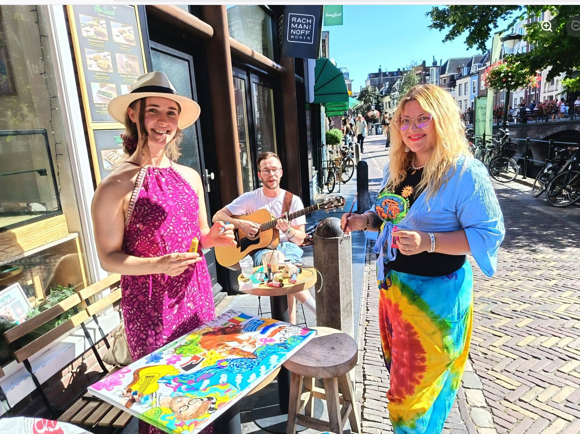The painters smiling by the canvas on the sunny street, with the guitarist playing behind them